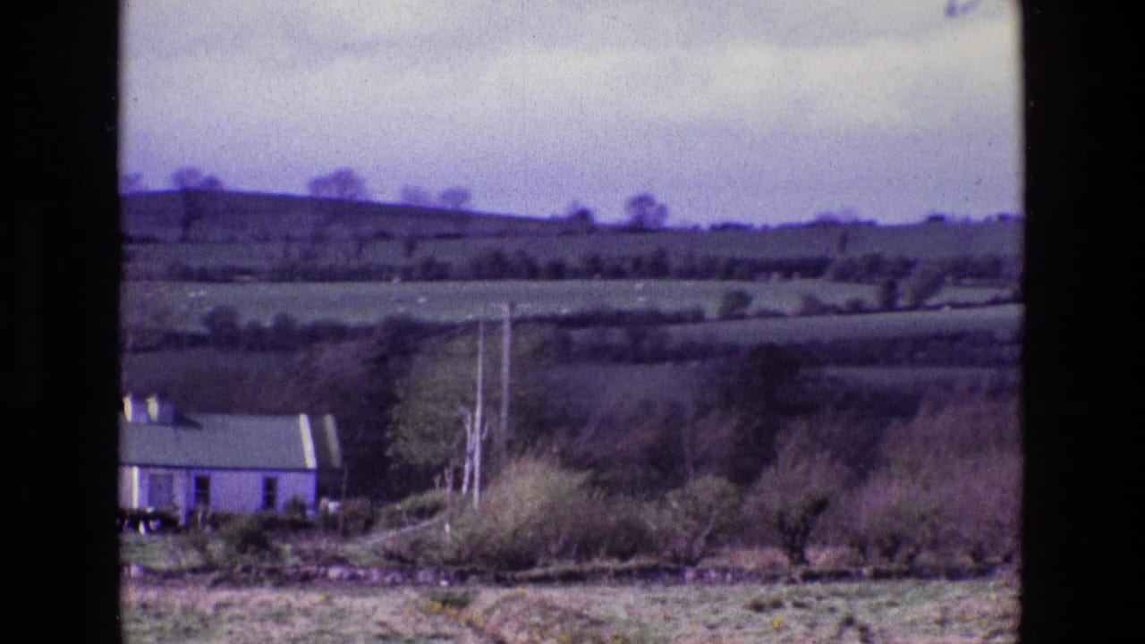 1969 Ireland home-movie frame showing rolling fields, hedgerows, utility poles, trees, and a small pale house at the lower left.
