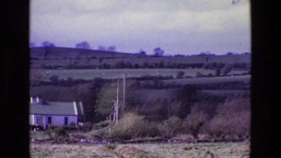 1969 Ireland home-movie frame showing rolling fields, hedgerows, utility poles, trees, and a small pale house at the lower left.