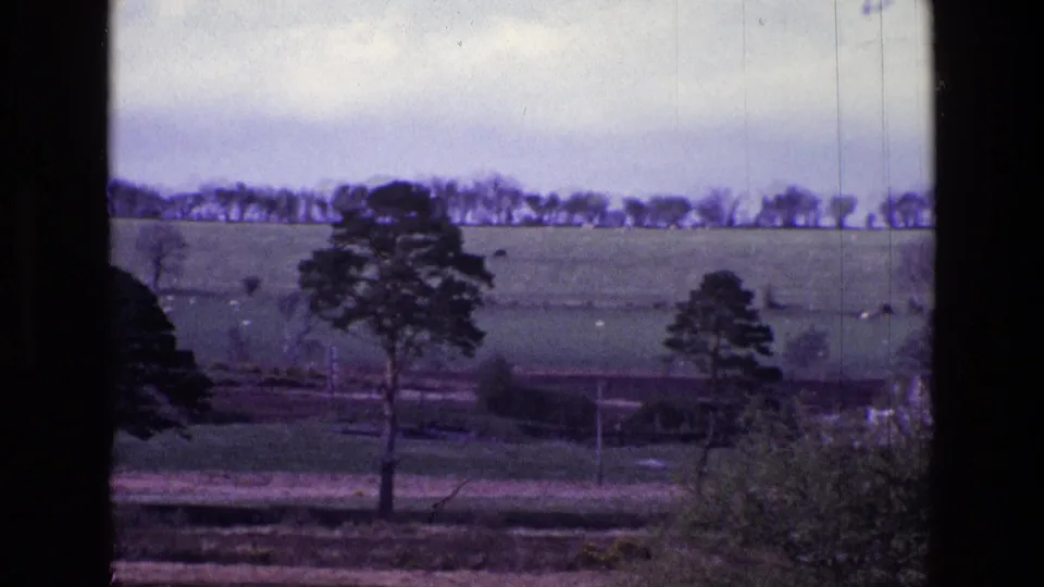 1969 Ireland 8mm frame showing rolling green fields, scattered trees, a distant tree line, cloudy sky, and visible film gate edges.