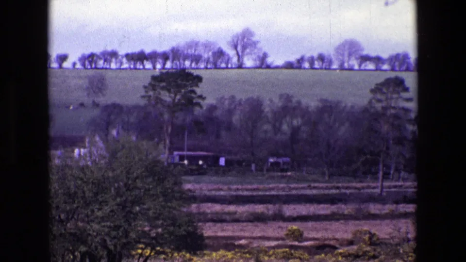 1969 Ireland 8mm view of fields, scattered trees, low rural buildings, foreground vegetation, and a cloudy sky.