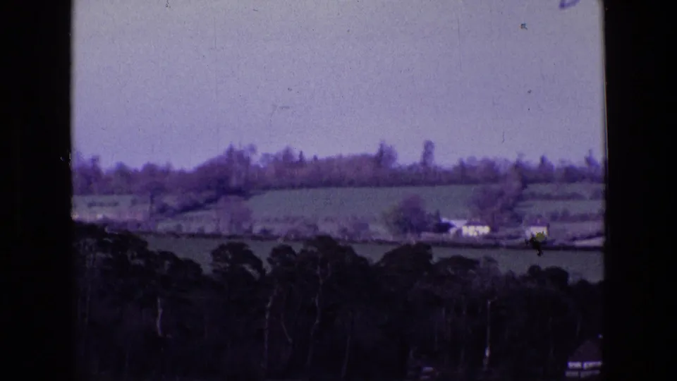 1969 8mm countryside frame showing distant white farm buildings, divided fields, hedgerows, and a dark tree foreground in Ireland.