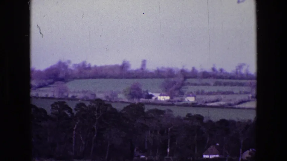 Rural Ireland in 1969, with rolling fields, small distant buildings, hedgerows, and dark trees along the lower frame.