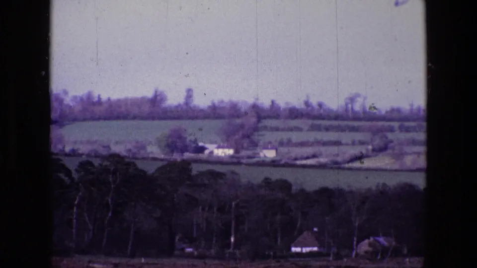 1969 Ireland 8mm frame showing green fields, hedgerows, wooded foreground, and small distant farm buildings under a pale sky.