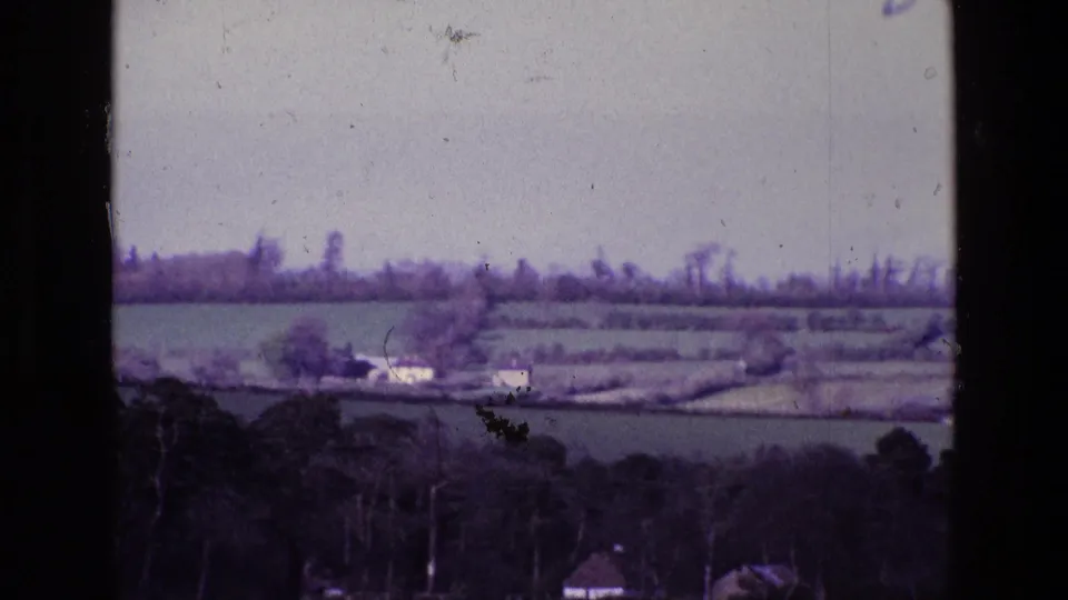1969 Ireland countryside frame with hedged fields, bare trees, small white buildings, and soft 8mm film borders.