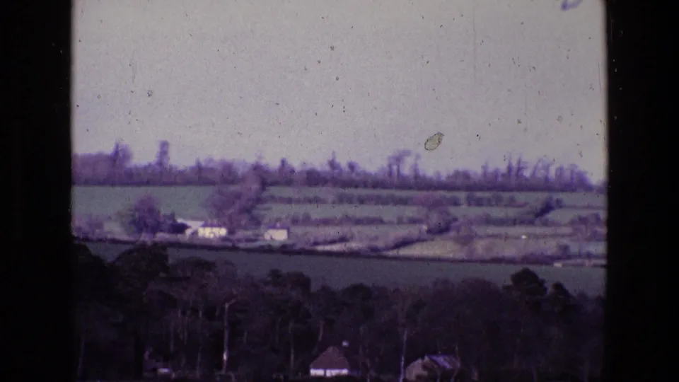 1969 Ireland 8mm frame with blurred pasture, hedgerows, scattered farm buildings, and wooded foreground from a moving viewpoint.