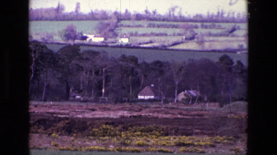 1969 Ireland countryside frame showing a small house, hedged fields, bare trees, and scrubby foreground with visible 8mm grain.