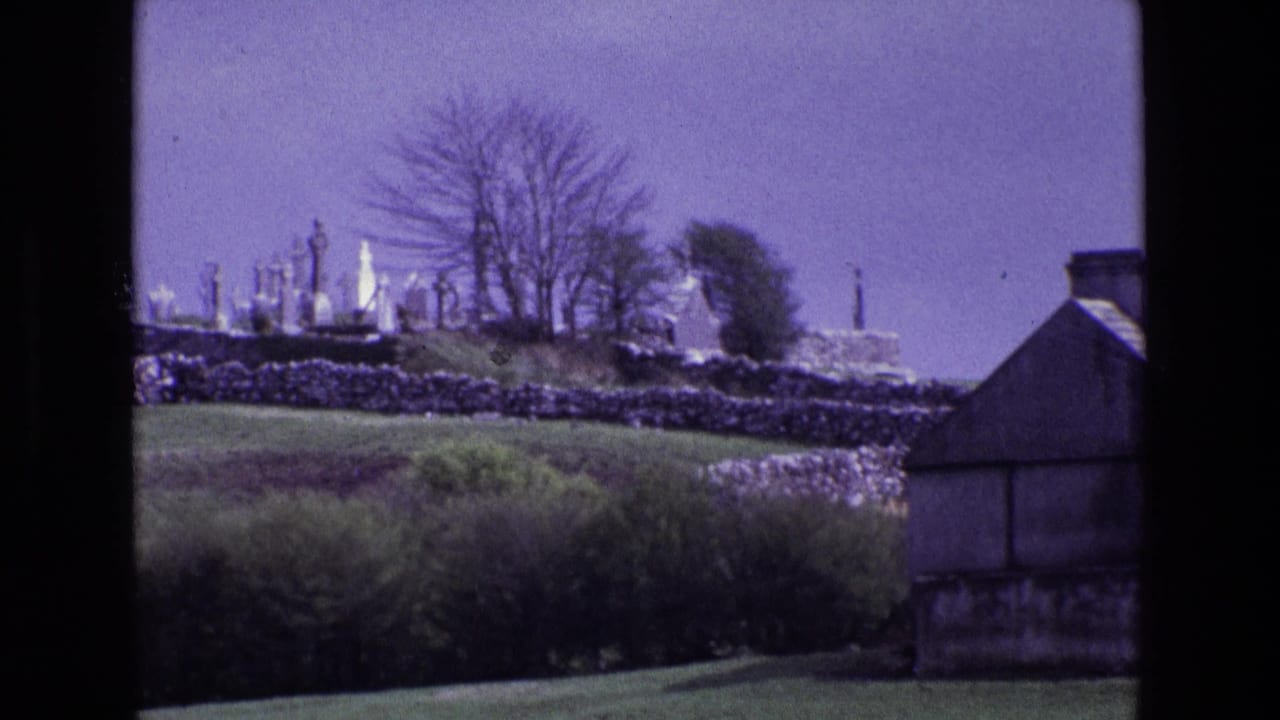 Hilltop cemetery beyond stone walls with trees, hedges, and a building edge.