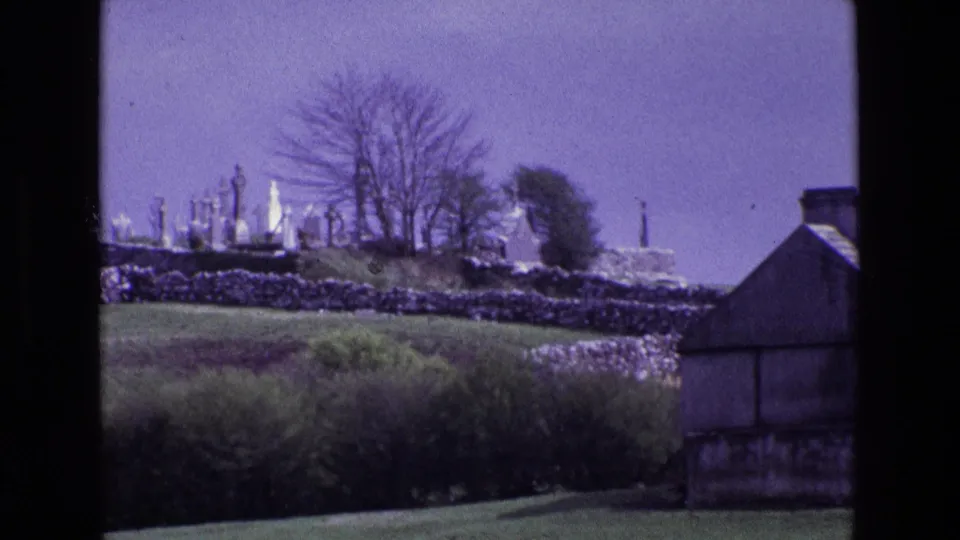 Irish rural hillside view showing a cemetery with upright gravestones behind stone walls, with trees and a building at frame right.