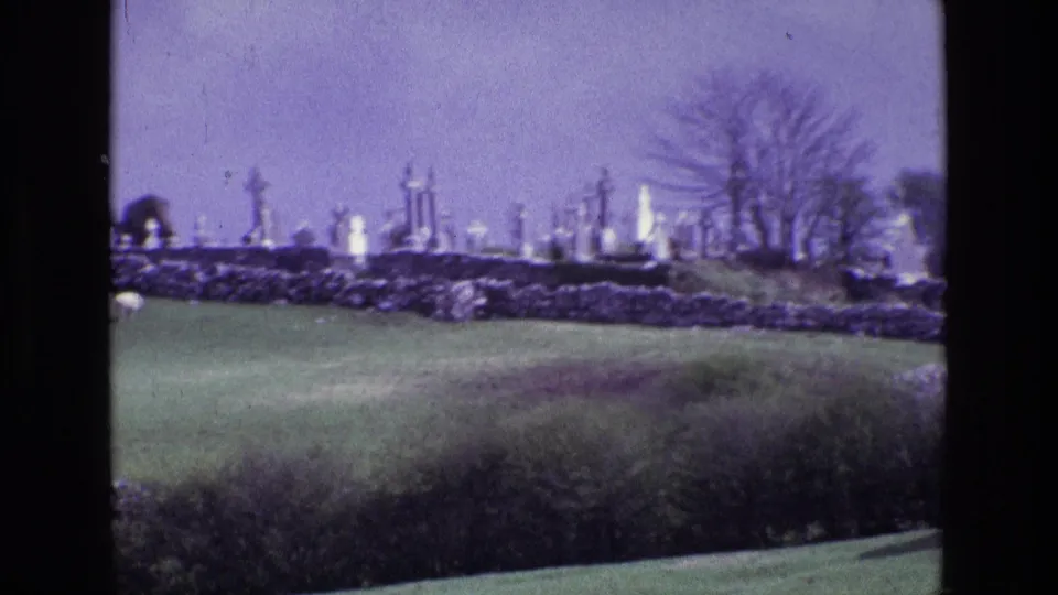 1969 Ireland countryside frame with a hilltop cemetery, pale gravestones, stone boundary walls, pasture, and hedgerow foreground.
