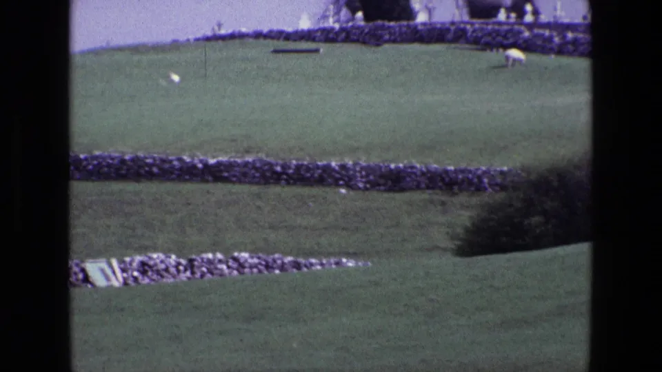 1969 Ireland home-movie frame showing a sloped green field, layered stone walls, a distant sheep, and soft 8mm grain.