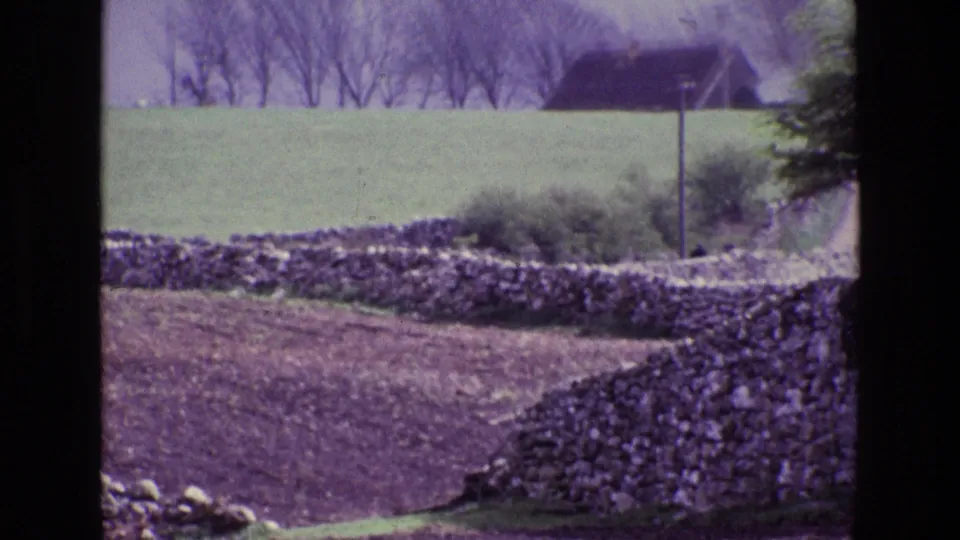 1969 Ireland home-movie view of dry-stone field boundaries, bare ground, pasture, and a distant small structure.