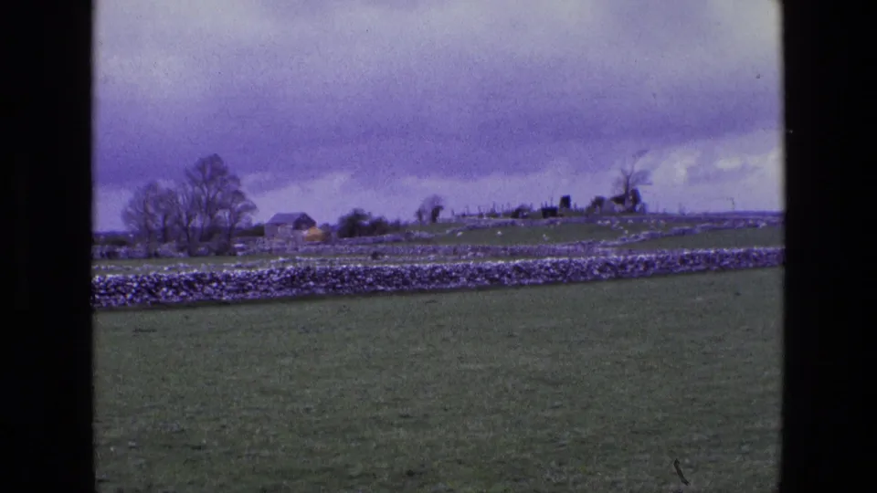 1969 home-movie footage of rural Ireland frames a green field, stone wall, trees, and small distant buildings under a moody sky.