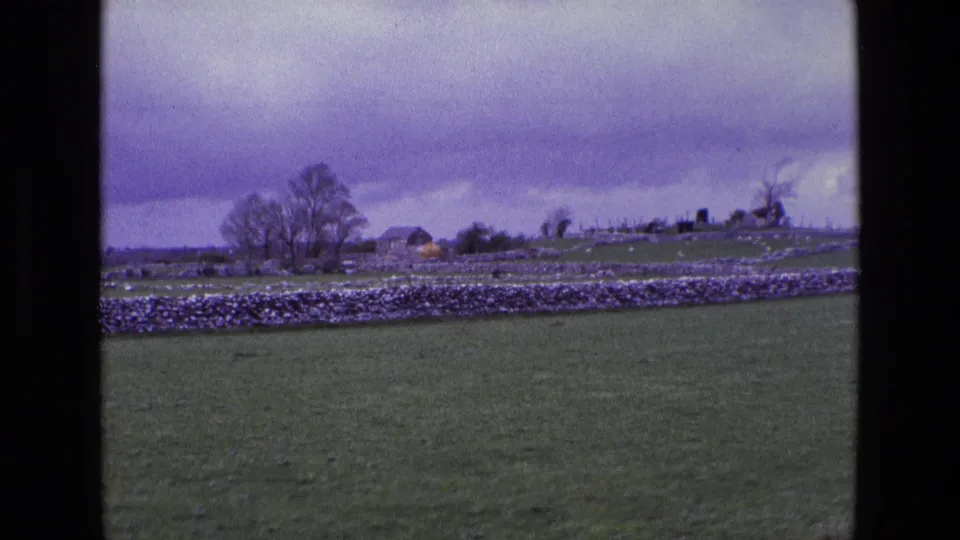 A rural Irish field in 1969 appears beyond the 8mm frame edge, with stone walls, trees, small buildings, and heavy cloud cover.