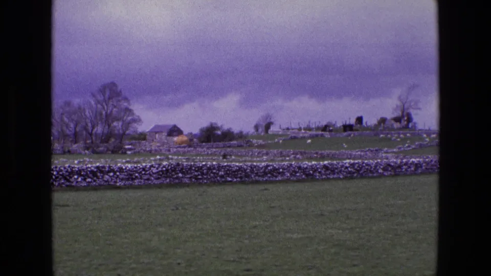 1969 8mm view of rural Ireland with stone-walled fields, distant farm buildings, trees, and cemetery monuments on a low hill.
