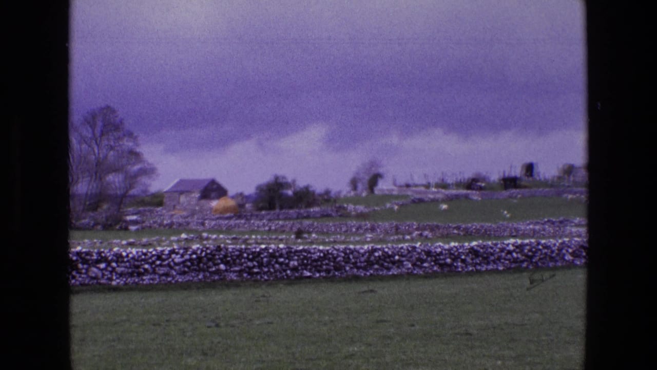1969 rural Ireland frame showing pasture, dry stone walls, a small outbuilding, hay-colored mound, trees, and cemetery markers beyond.