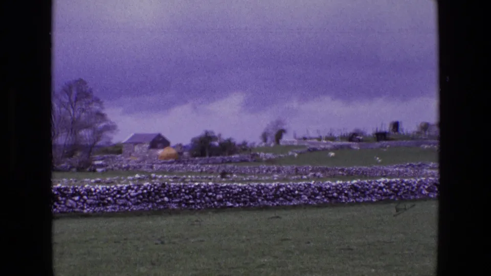 1969 rural Ireland frame showing pasture, dry stone walls, a small outbuilding, hay-colored mound, trees, and cemetery markers beyond.