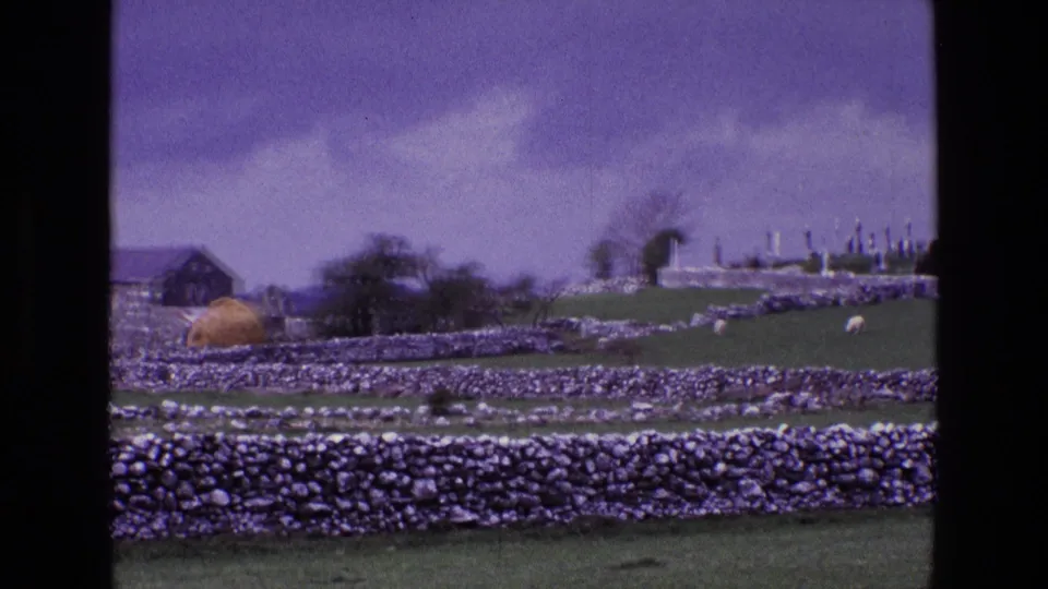 1969 8mm countryside view in Ireland with layered stone walls, green fields, a barn-like structure, hay-colored mound, trees, and cemetery markers.