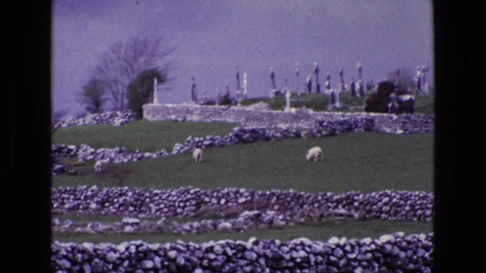 1969 Irish countryside frame with sheep grazing below dry stone walls and a hillside cemetery lined with upright monuments.