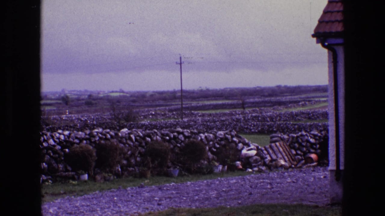 Rural Irish landscape in 1969 with stone field walls, a gravel foreground, utility pole, roof edge, and small debris near the wall.