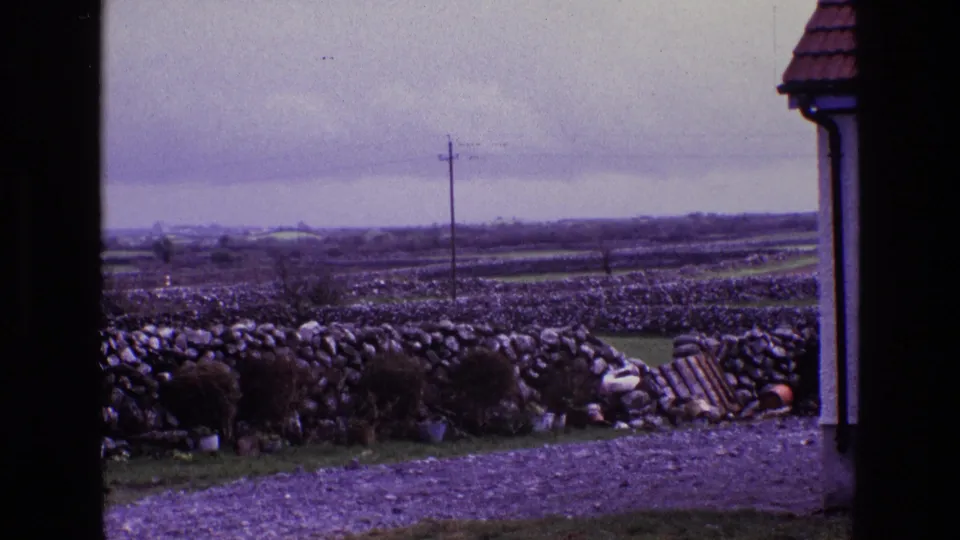 Rural Irish landscape in 1969 with stone field walls, a gravel foreground, utility pole, roof edge, and small debris near the wall.