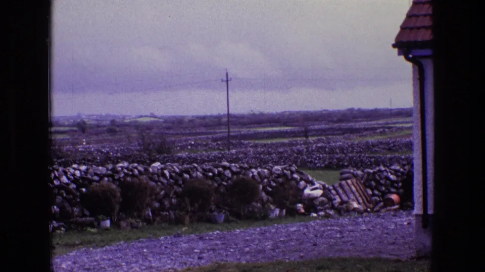 1969 Ireland countryside frame showing a dark doorway, gravel ground, potted plants, dry-stone walls, fields, and a utility pole.