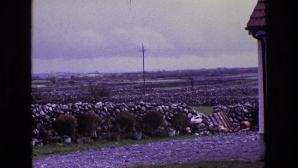 1969 rural Ireland view with layered fields behind stone walls, a gravel foreground, utility pole, doorway shadow, and roof edge at right.