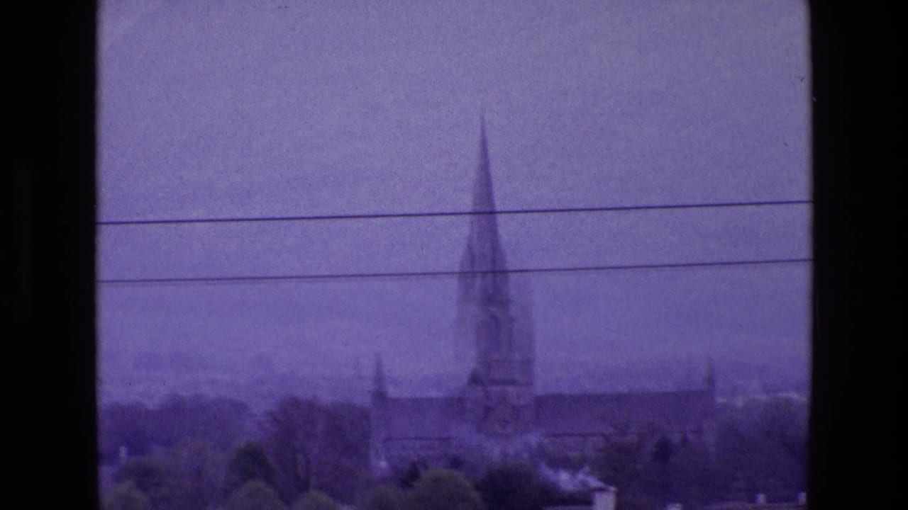 Distant Irish church with a tall spire, trees, rooftops, and utility lines in a purple-tinted 8mm frame.