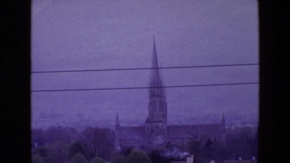 Purple-cast 8mm view of a church spire and roofline in Ireland, with trees and overhead wires in the foreground.