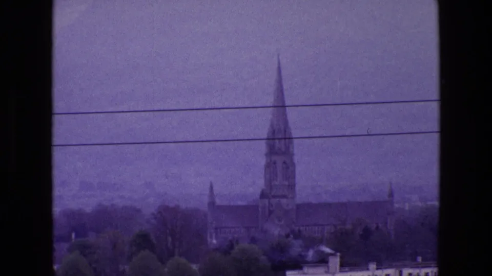 Distant 1969 Ireland home-movie frame showing a church exterior with a tall spire among trees and rooftops.