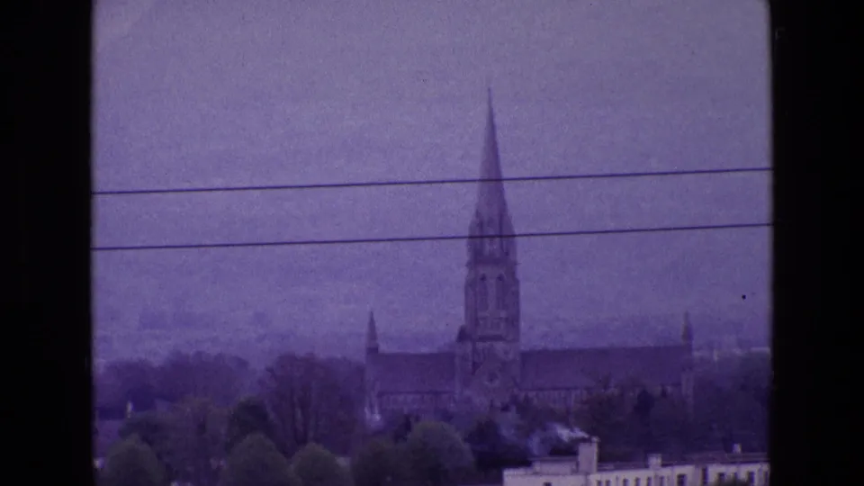 1969 Ireland 8mm frame showing a tall church spire above rooftops, crossed by overhead wires under a hazy sky.