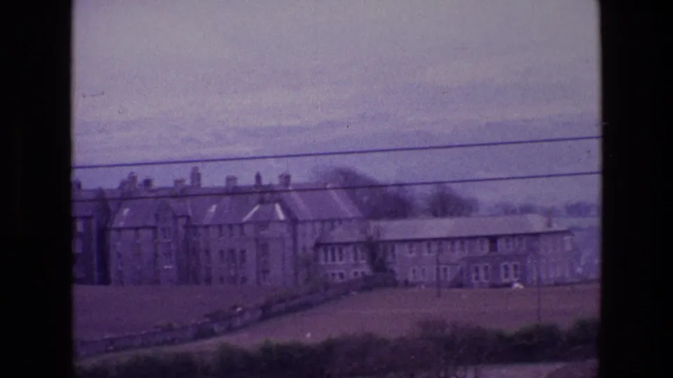 1969 Ireland 8mm frame showing stone houses on a hillside, open ground, overhead wires, and a pale sky.