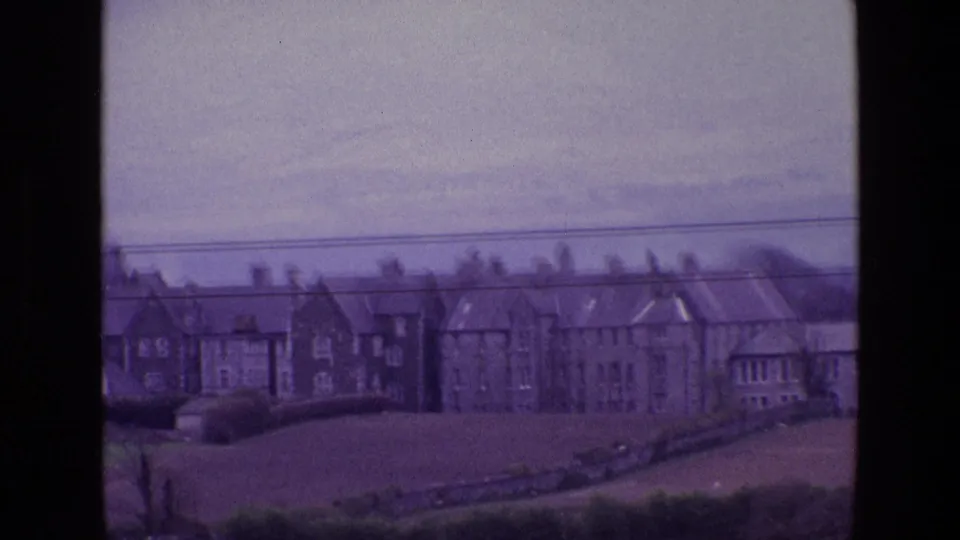1969 Ireland 8mm frame showing a row of rural houses viewed across sloping pasture with hedges and power lines.
