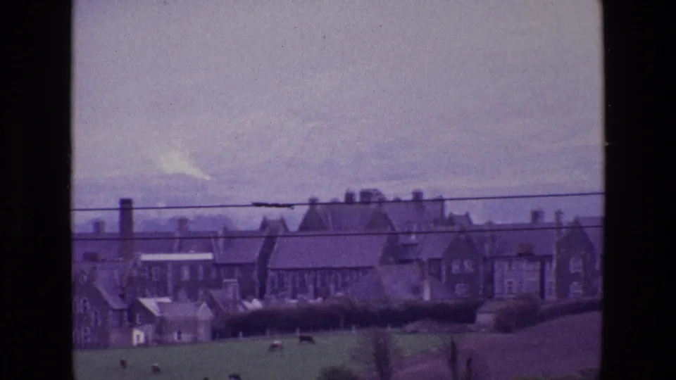 A soft 8mm frame from 1969 Ireland showing cows grazing before rural houses, hedges, rooftops, and a cloudy sky.