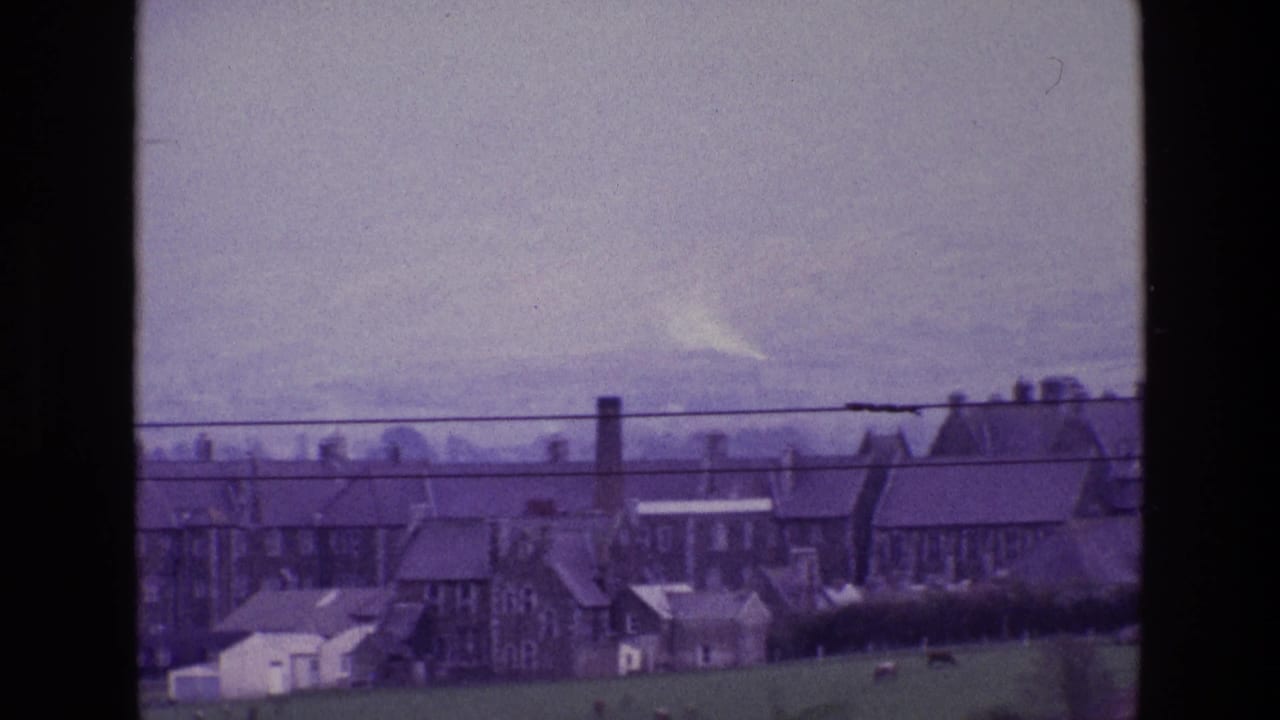 1969 Ireland home-movie frame with clustered stone buildings, chimneys, grazing field, cattle, and utility wires across the foreground view.