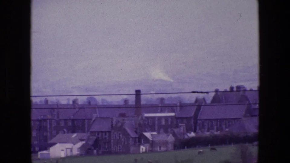 1969 Ireland home-movie frame with clustered stone buildings, chimneys, grazing field, cattle, and utility wires across the foreground view.