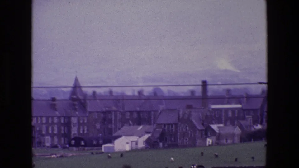 1969 Ireland 8mm frame showing a pointed tower or spire, stone buildings, slate roofs, pasture, cattle, and a pale overcast sky.