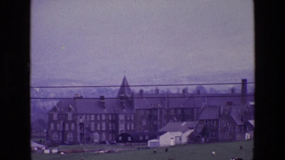 1969 Ireland home-movie frame with a distant stone town, pointed tower, chimneys, green field, grazing cattle, and overhead wires.