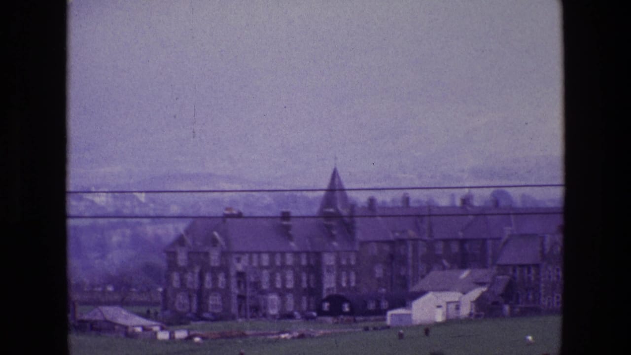 1969 Ireland home-movie frame showing a distant stone house with a pointed tower, green fields, hazy mountains, and power lines.