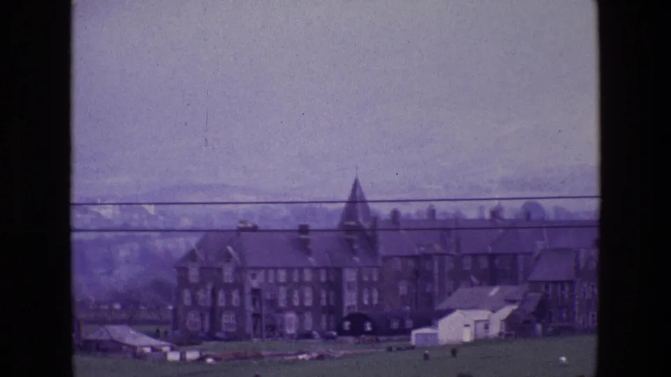 1969 Ireland home-movie frame showing a distant stone house with a pointed tower, green fields, hazy mountains, and power lines.