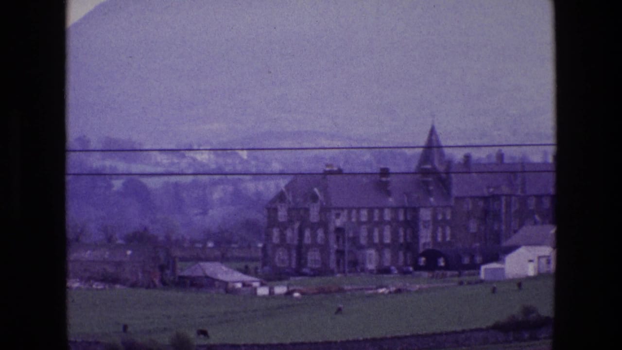 Distant 1969 Ireland view of a multi-story stone house with tower, farm structures, green pasture, mountains, and wires across the frame.