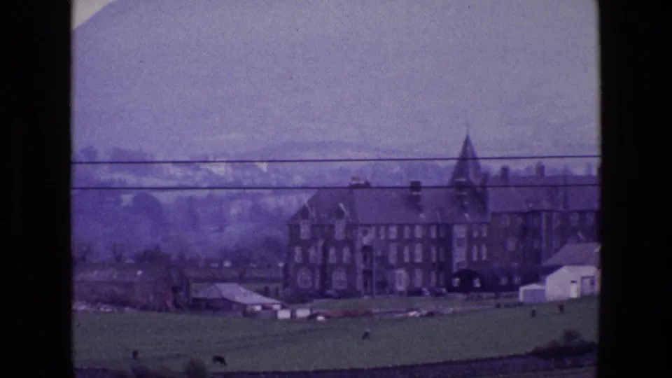 Distant 1969 Ireland view of a multi-story stone house with tower, farm structures, green pasture, mountains, and wires across the frame.