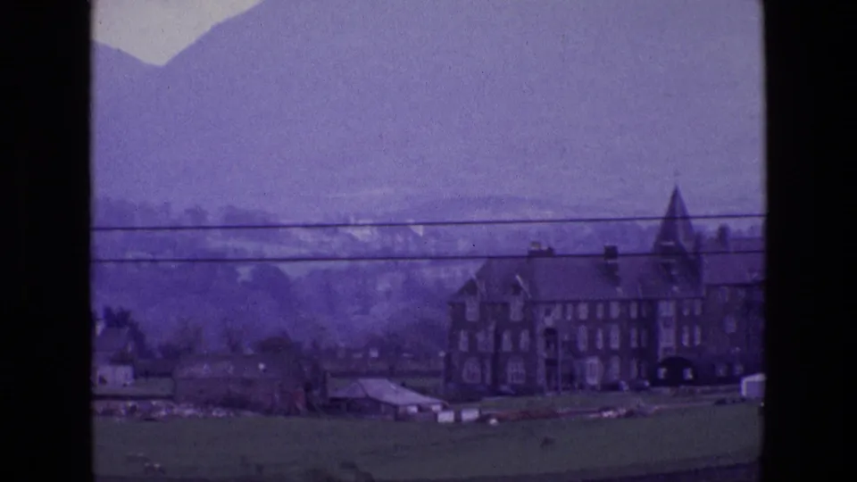 1969 Ireland frame with the large stone house shifted to the right, set among fields, sheds, mountain haze, and overhead utility lines.