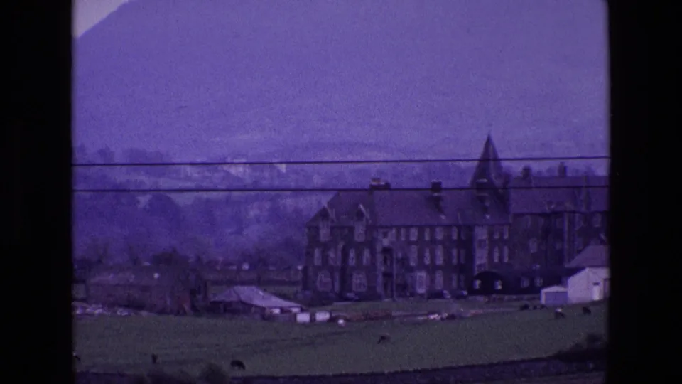 Final 1969 Ireland frame showing the house partly to the right, with green fields, outbuildings, mountains, haze, and visible film grain.