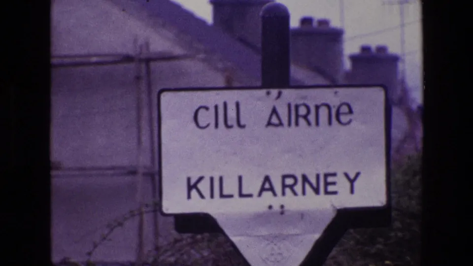 Vintage 8mm view of the bilingual Killarney sign, with rooflines, chimneys, and soft purple color shift in the background.