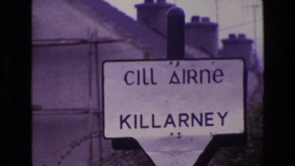 1969 Ireland home-movie frame holding on the Killarney place sign, with chimneys and a distant hillside beyond the rooftops.
