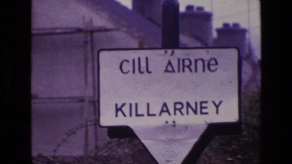1969 Ireland home-movie frame showing a bilingual Cill Airne Killarney road sign behind foreground barbed wire.