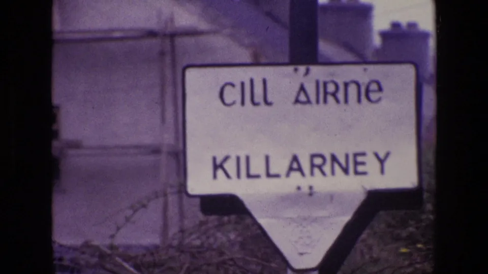1969 Ireland frame of a white Cill Airne Killarney sign, softly focused with barbed wire crossing the foreground.