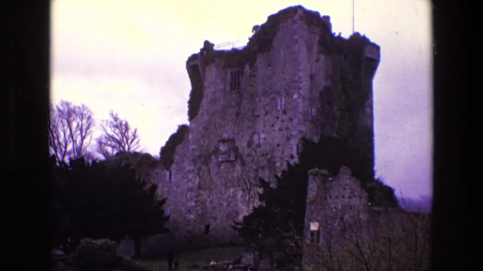 1969 Ireland home-movie frame showing a weathered stone castle-like ruin with ivy, trees, and overcast sky.