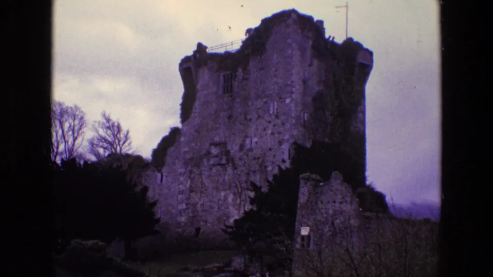 1969 Ireland 8mm frame of a crumbling stone tower ruin, ivy-covered sections, leafless trees, and overcast sky.