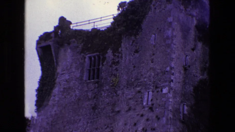 1969 Ireland 8mm frame showing a weathered stone ruin with ivy, a barred window, roof railing, and dark film gate edges.